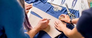 Cutting blue fabric on the table full of tailoring tools. Close-up view on the hands, fabric and fashion drawings