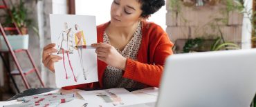 Young attractive woman with dark curly hair sitting at the table dreamily showing fashion illustrations in laptop spending time in modern cozy workshop with big windows